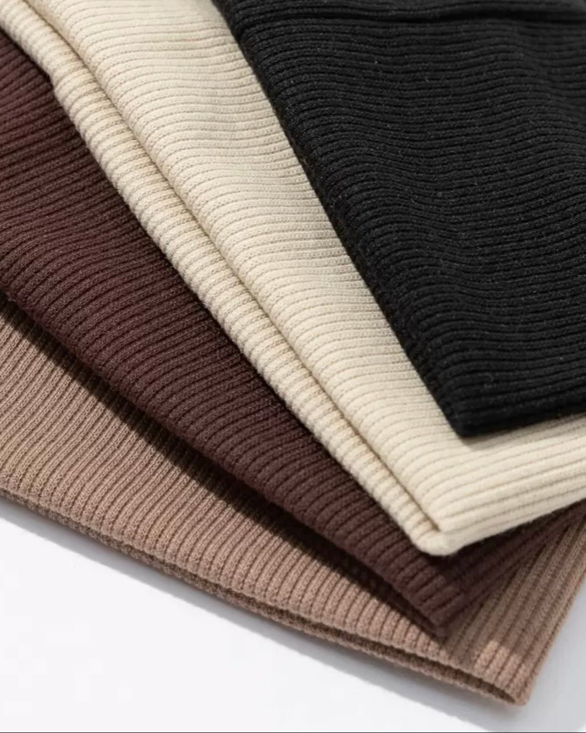 Stack of folded ribbed knit sweaters in black, beige, and brown on a white background